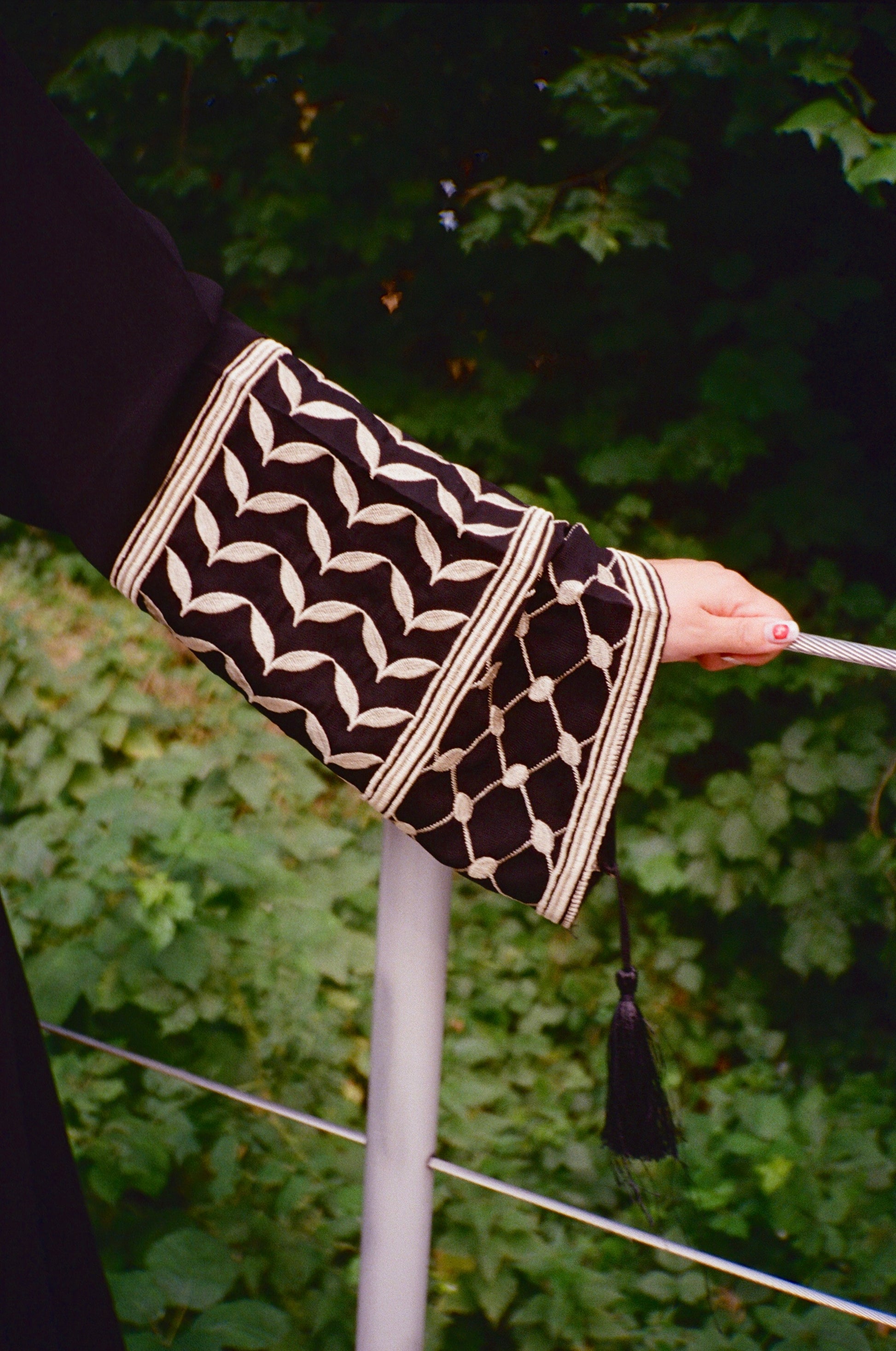 Black and white patterned keffiyeh sleeve with a tassel held by a hand against a green leafy background