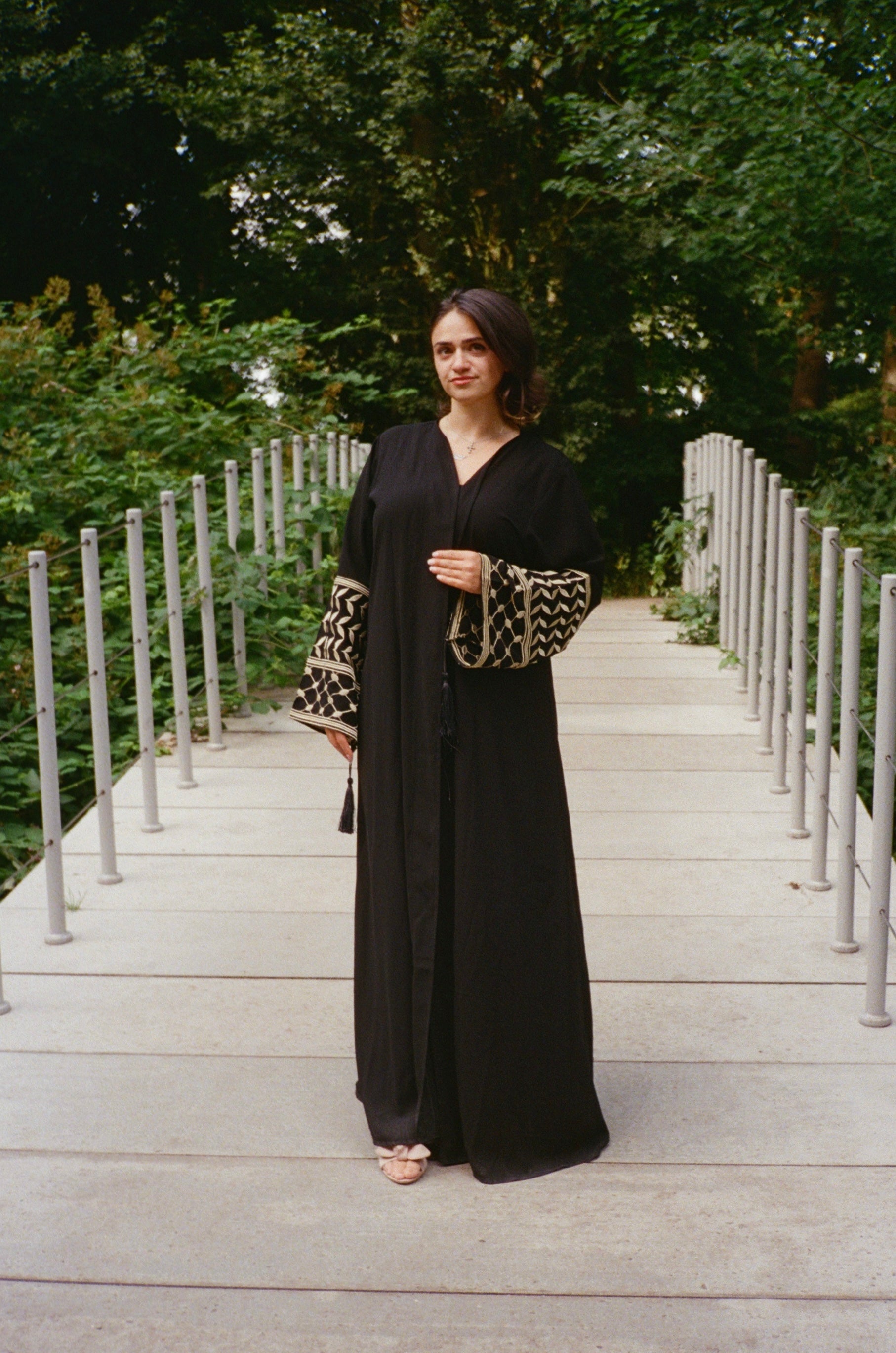 Woman in a black thobe with a keffiyeh print sleeve standing on a wooden bridge with greenery in the background