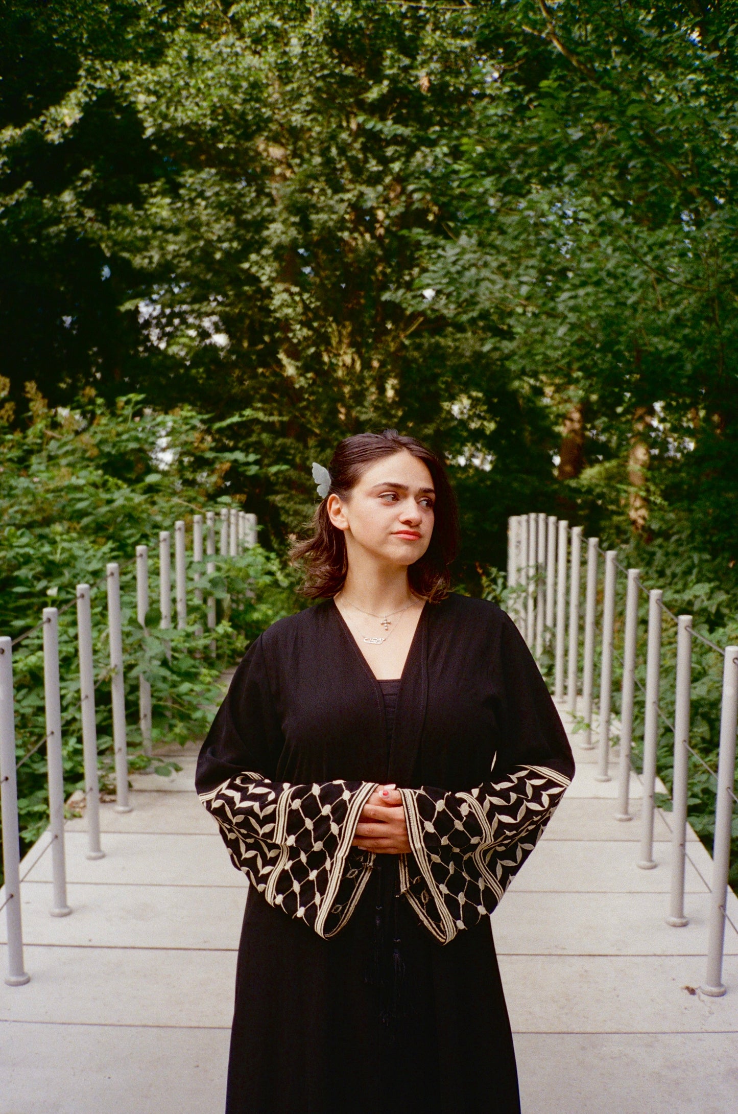 Woman in a black thobe with a patterned keffiyeh sleeves standing outdoors with greenery in the background