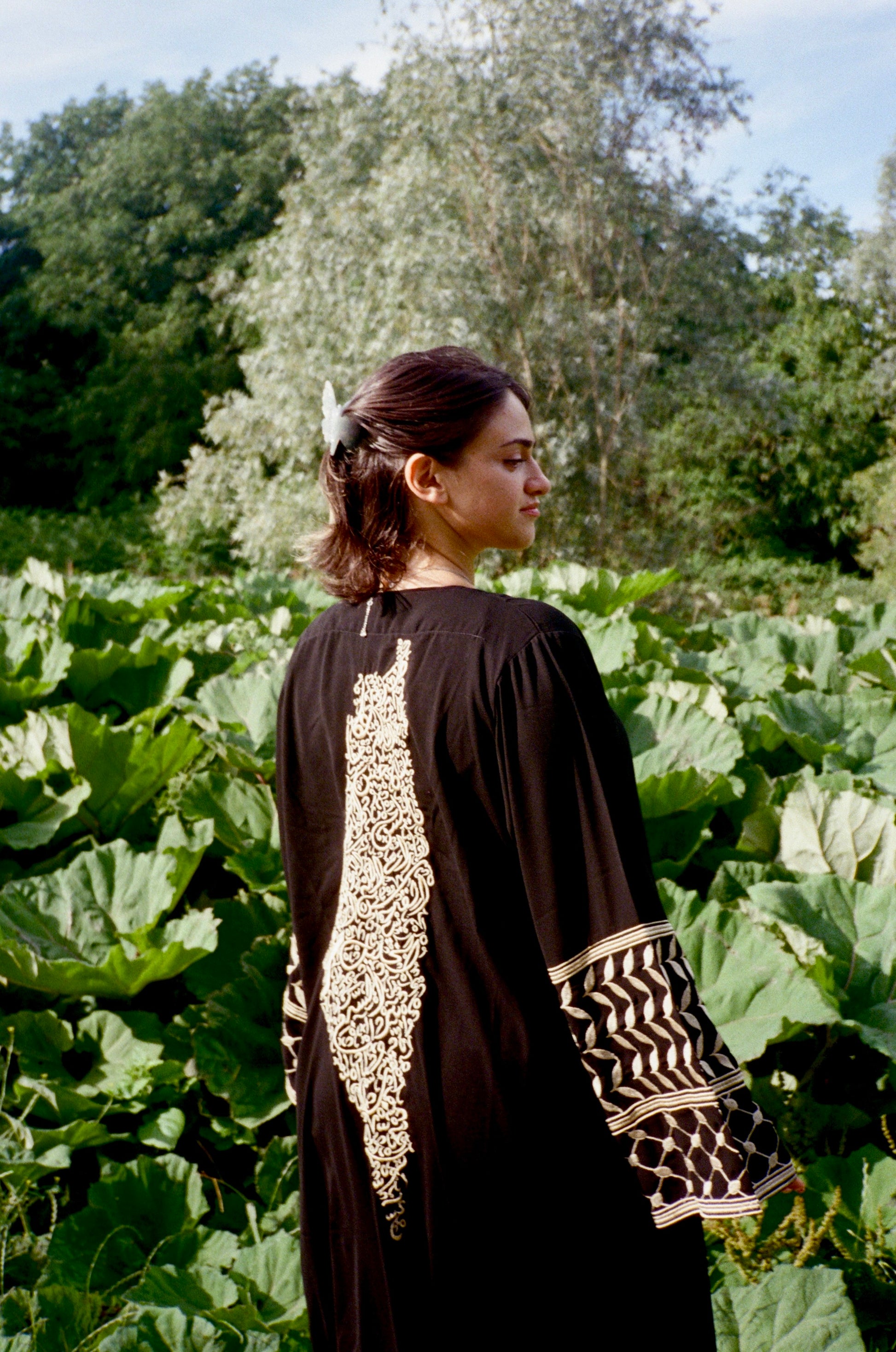 Woman in a black thobe with Palestine map embroidered dress standing in a field of green plants