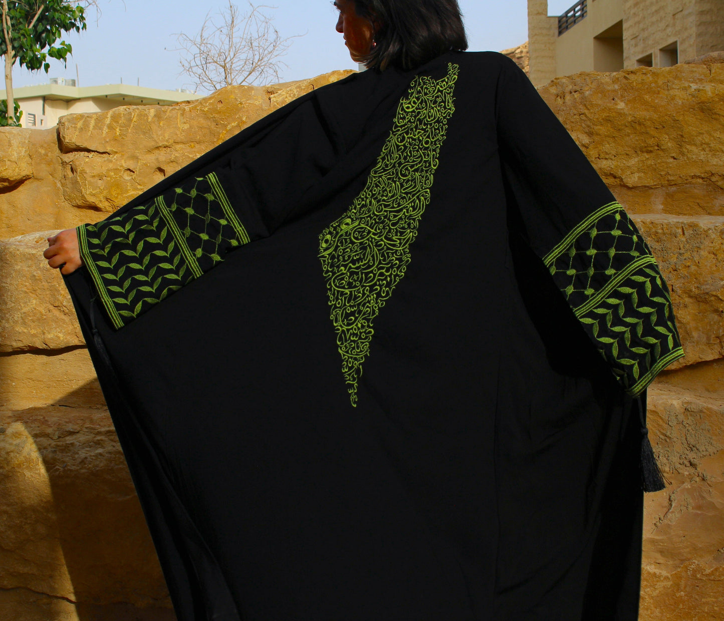 Person wearing a black garment with green patterns palestine map and keffiyeh sleeves against a stone wall