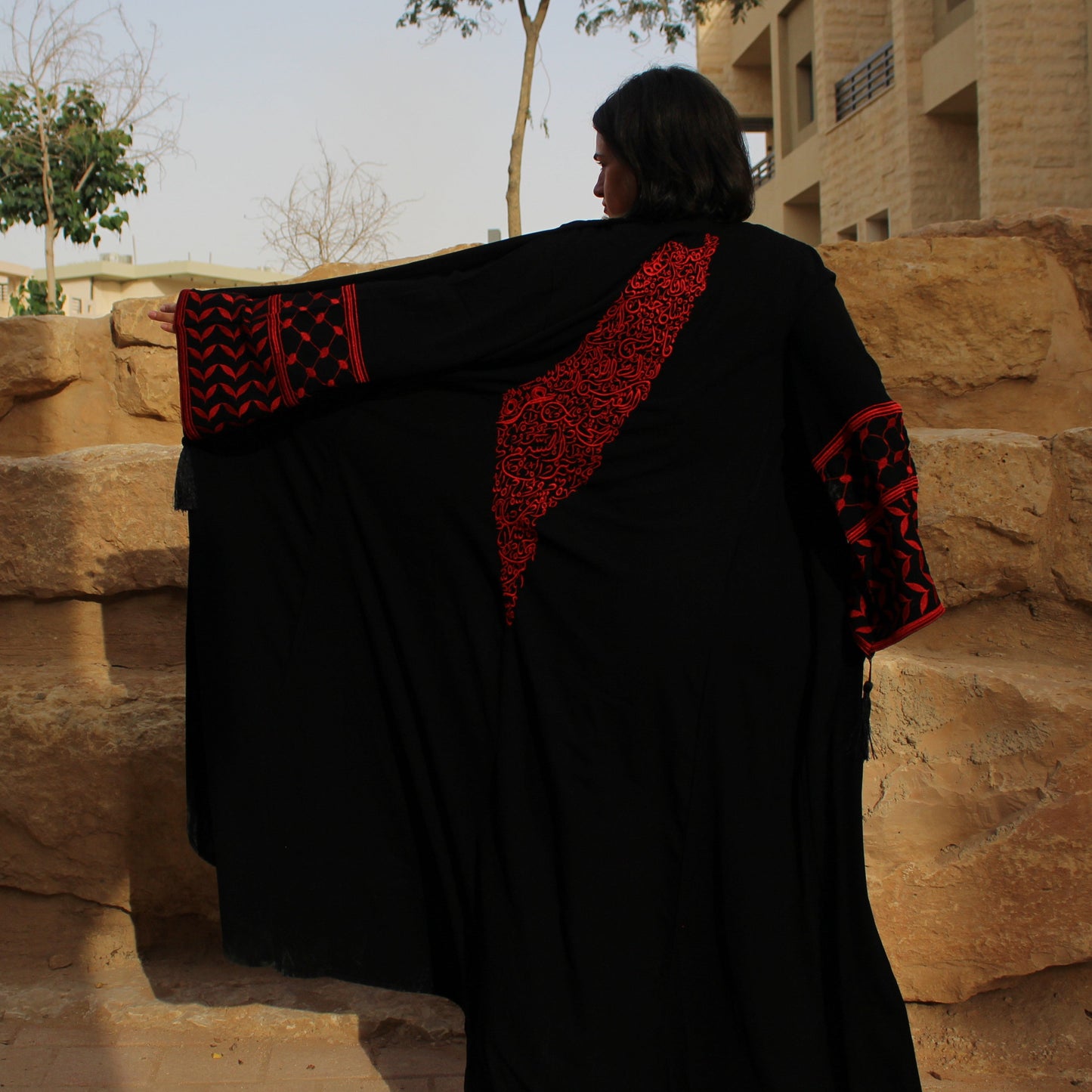 Person wearing a black and red palestine map traditional garment standing in front of stone steps and buildings.