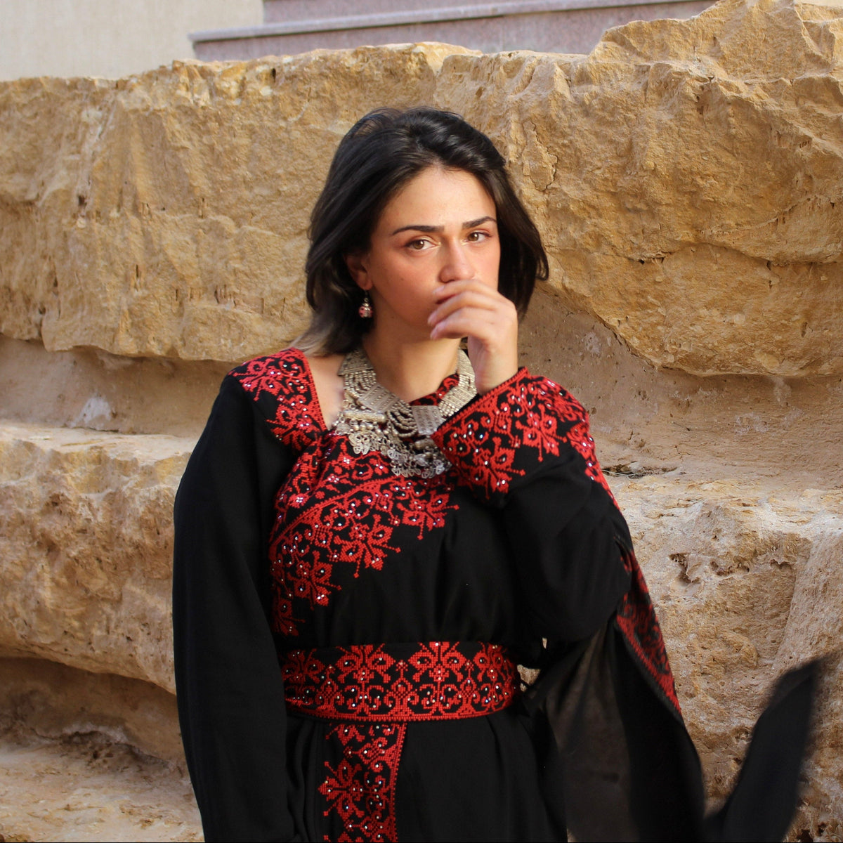 A woman wearing a black traditional dress with red floral embroidery and a silver necklace, standing in front of a large tan stone wall.