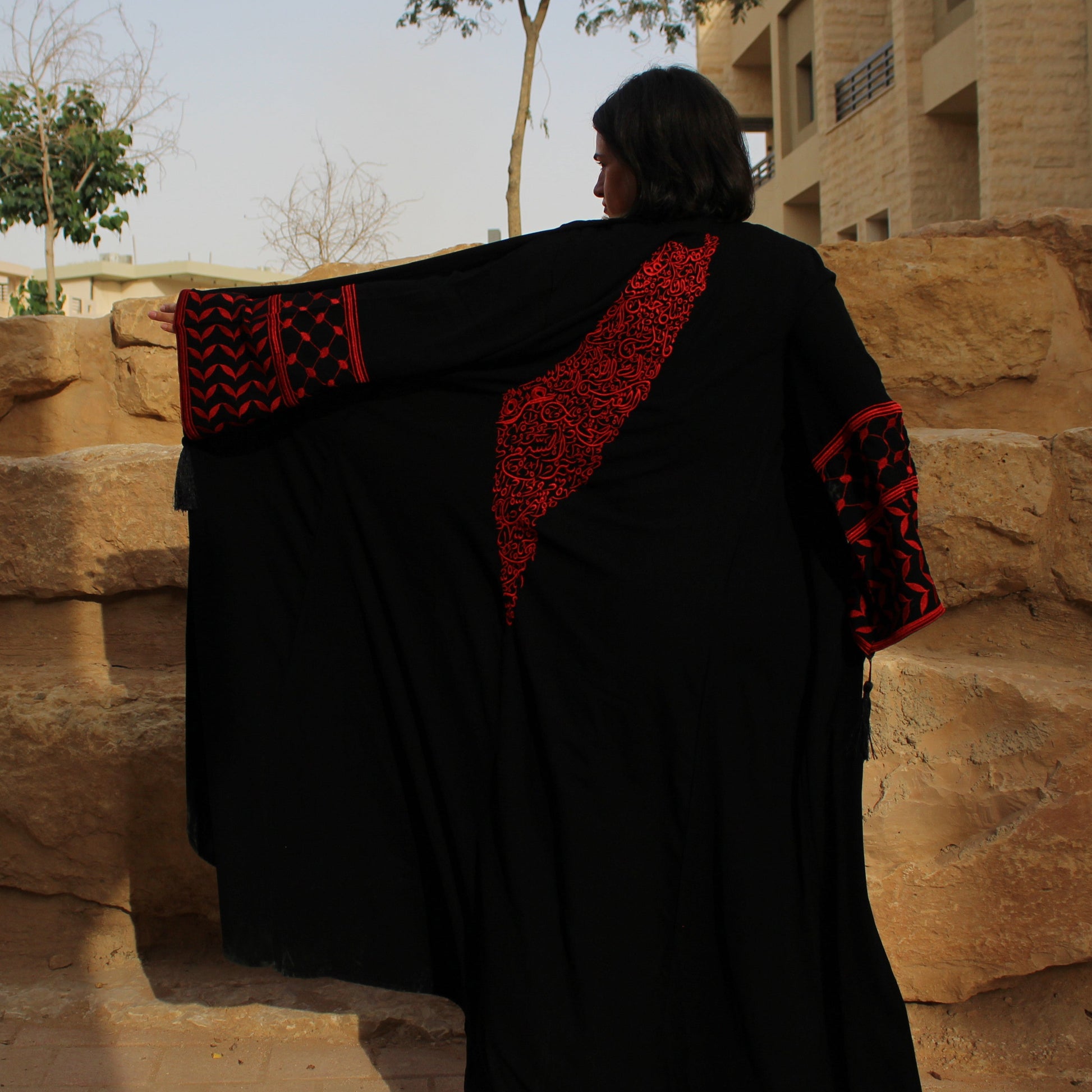 Person wearing a black and red palestine map traditional garment standing in front of stone steps and buildings.