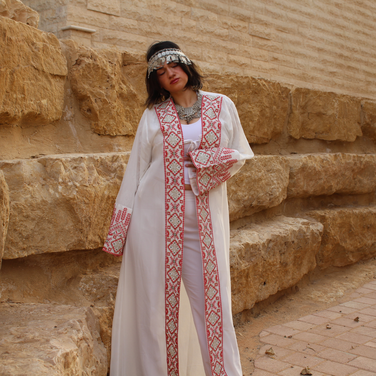 Woman in a long, white dress with tatreez red patterns standing in front of stone steps.