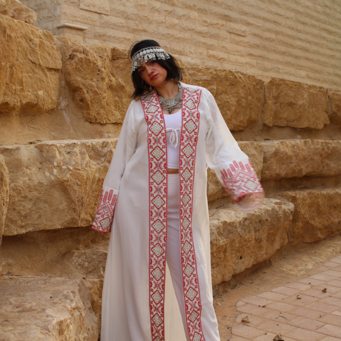 Person wearing a traditional outfit with tatreez  intricate patterns in front of a stone wall.